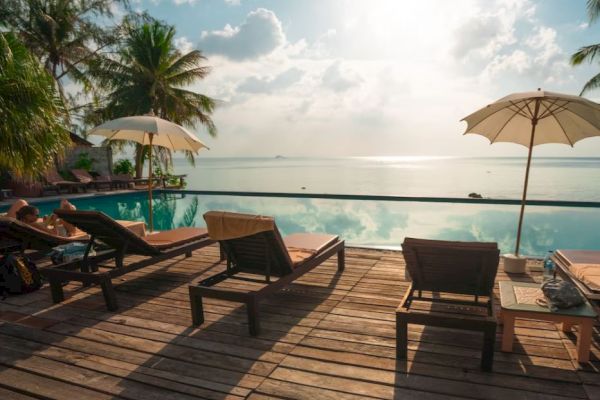 A serene poolside scene with lounge chairs, umbrellas, and a view of the ocean, framed by palm trees, under a partly cloudy sky.