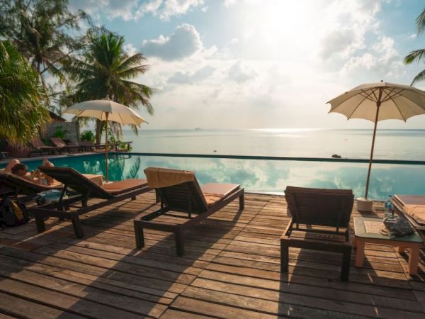 A serene poolside scene with lounge chairs, umbrellas, and a view of the ocean, framed by palm trees, under a partly cloudy sky.