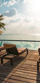 The image shows a tropical poolside scene with sun loungers, umbrellas, and palm trees, overlooking a serene body of water under a partly cloudy sky.