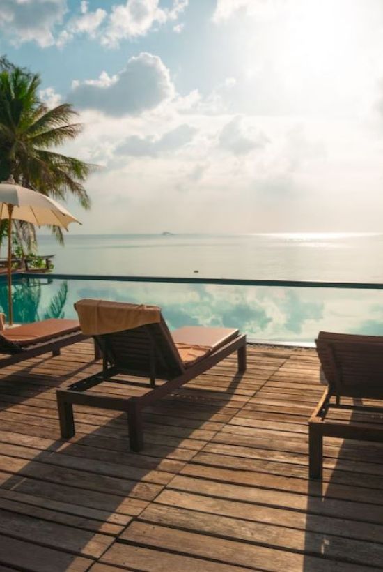 The image shows a serene poolside setup with lounge chairs and umbrellas overlooking an infinity pool and a scenic body of water under a partly cloudy sky.