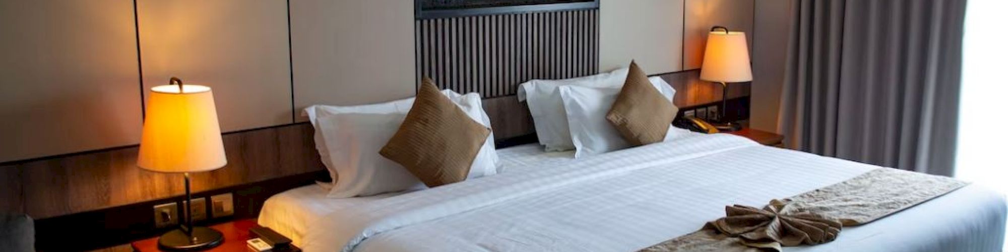 A neatly made bed in a well-lit hotel room with bedside lamps, cushions, and a small towel decoration at the foot of the bed.