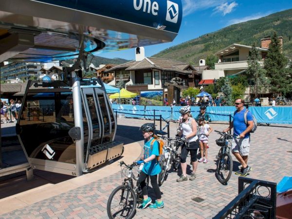 A Vail bike event: riders line up with bicycles beside an open lift/gondola in a mountain town, under a blue &ldquo;One&rdquo; sign.