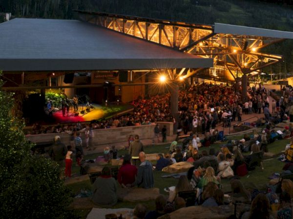 An outdoor evening concert at an amphitheater with a stage under large steel trusses, bright lights, and a full crowd seated and standing.