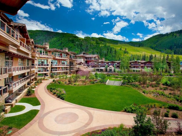 Scenic resort complex with balconies beside a manicured garden and winding walkway, set against green mountains under blue skies.