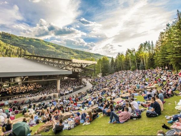 A packed outdoor amphitheater with people picnicking on the lawn beside a stage, surrounded by forested mountain hills.