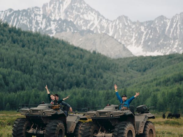 Two people riding ATVs with arms up, cruising through a green valley with snowy mountains in the background.
