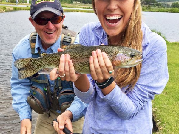 Smiling anglers pose with a speckled trout by a lake in the mountains during a fishing trip.