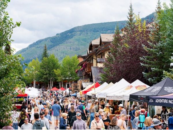 A busy outdoor festival in a mountain village, with many people, colorful tents, and shops beside forested hills.