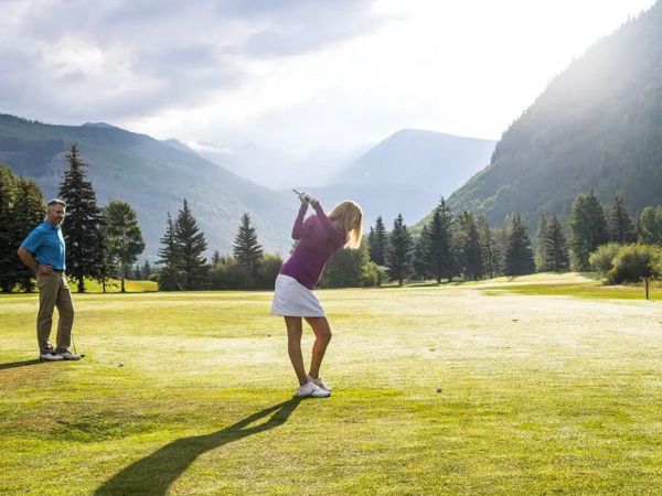 Golfer swings on a sunny mountain golf course with a man watching, surrounded by pine trees and misty hills.