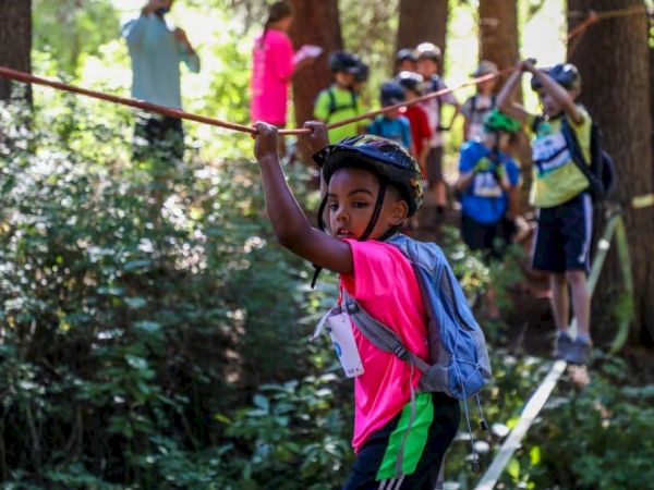 A child in a pink shirt and helmet leads a zipline-style forest obstacle course, with other kids watching behind the rope.