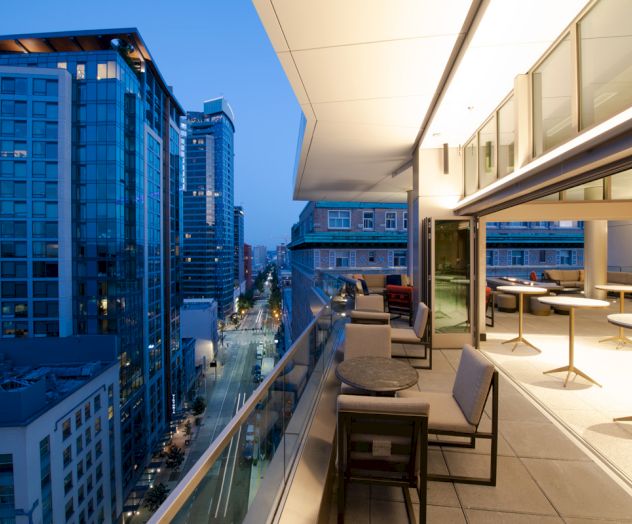 This image shows a modern rooftop terrace with seating, overlooking a cityscape with tall buildings at dusk or early evening.