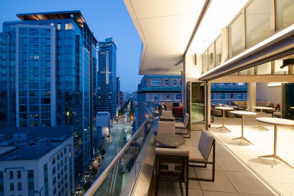 The image shows a balcony with chairs and tables overlooking a cityscape filled with modern high-rise buildings at dusk.