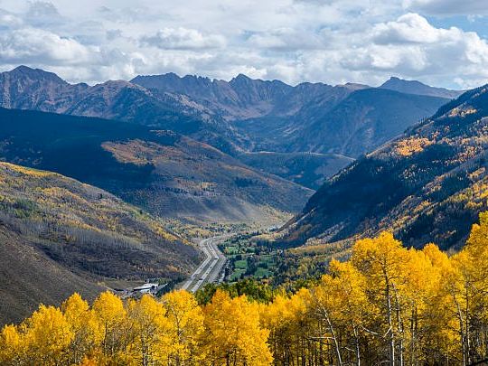 Autumn mountains unfold over a green valley with a winding road, framed by bright yellow trees under dramatic clouds.