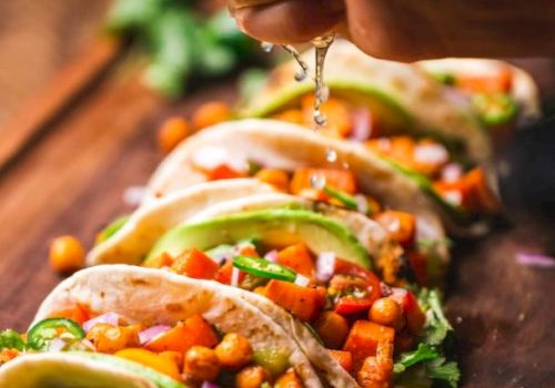 The image shows a hand squeezing lime over a row of chickpea and vegetable tacos garnished with avocado and cilantro on a wooden board.