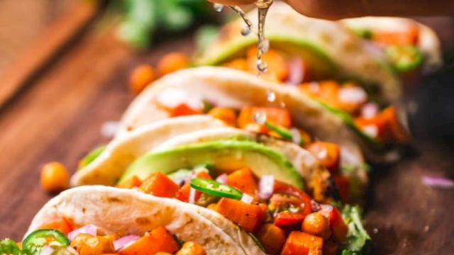 The image shows a hand squeezing lime over a row of chickpea and vegetable tacos garnished with avocado and cilantro on a wooden board.