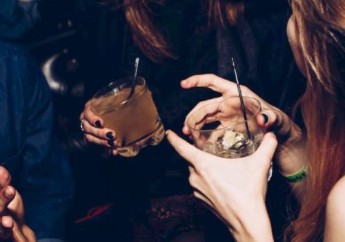 The image shows a group of people socializing, holding drinks, and having a lively conversation in what appears to be a club or party setting.