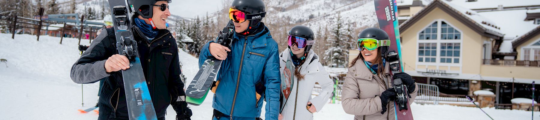 Four friends in ski gear walk through fresh snow, each carrying skis, smiling as they head to the slopes at a cozy mountain lodge.