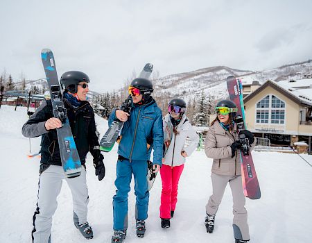 Four friends in ski gear walk through fresh snow, each carrying skis, smiling as they head to the slopes at a cozy mountain lodge.