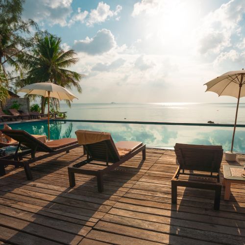 Sun loungers and umbrellas by a serene swimming pool, overlooking the ocean with a bright sun and some clouds in the sky.