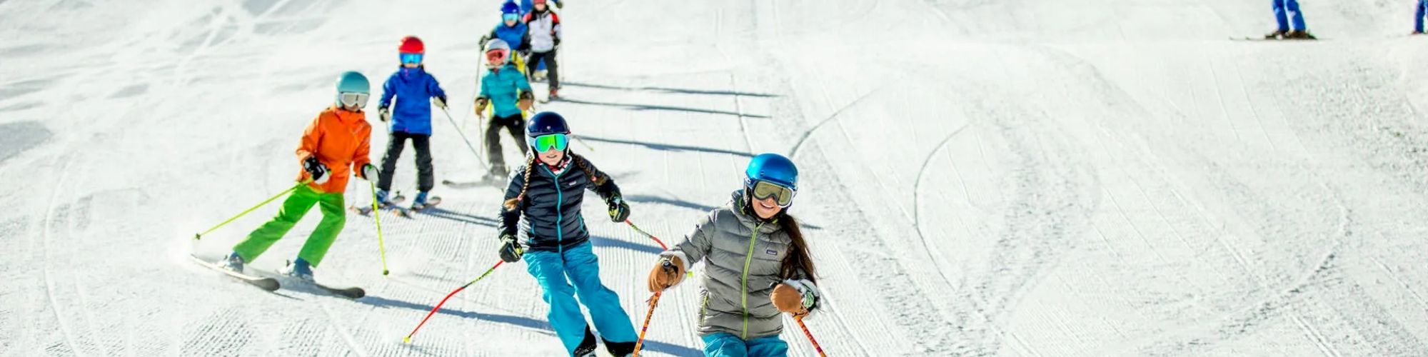 A group of children is skiing down a snowy slope, supervised by an adult. Each child is wearing ski gear, including helmets and poles.