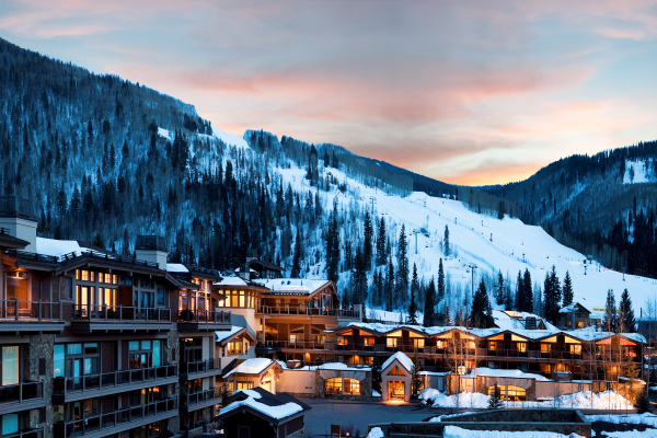 A snowy mountain resort at dusk with lit buildings below and ski slopes visible on the hills, under a colorful sky.