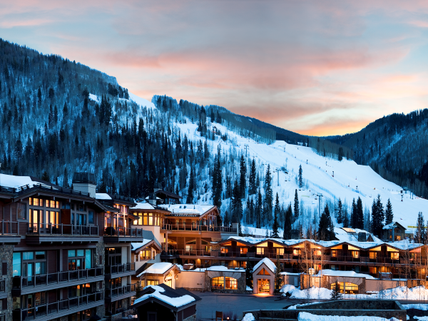 A snowy mountain resort at dusk with lit buildings below and ski slopes visible on the hills, under a colorful sky.