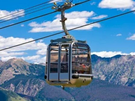 The image shows a cable car suspended on a cable with mountainous scenery in the background under a clear blue sky with some clouds.