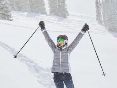 A person in winter gear stands in the snow, holding ski poles in the air with both arms raised, appearing to celebrate or pose happily.