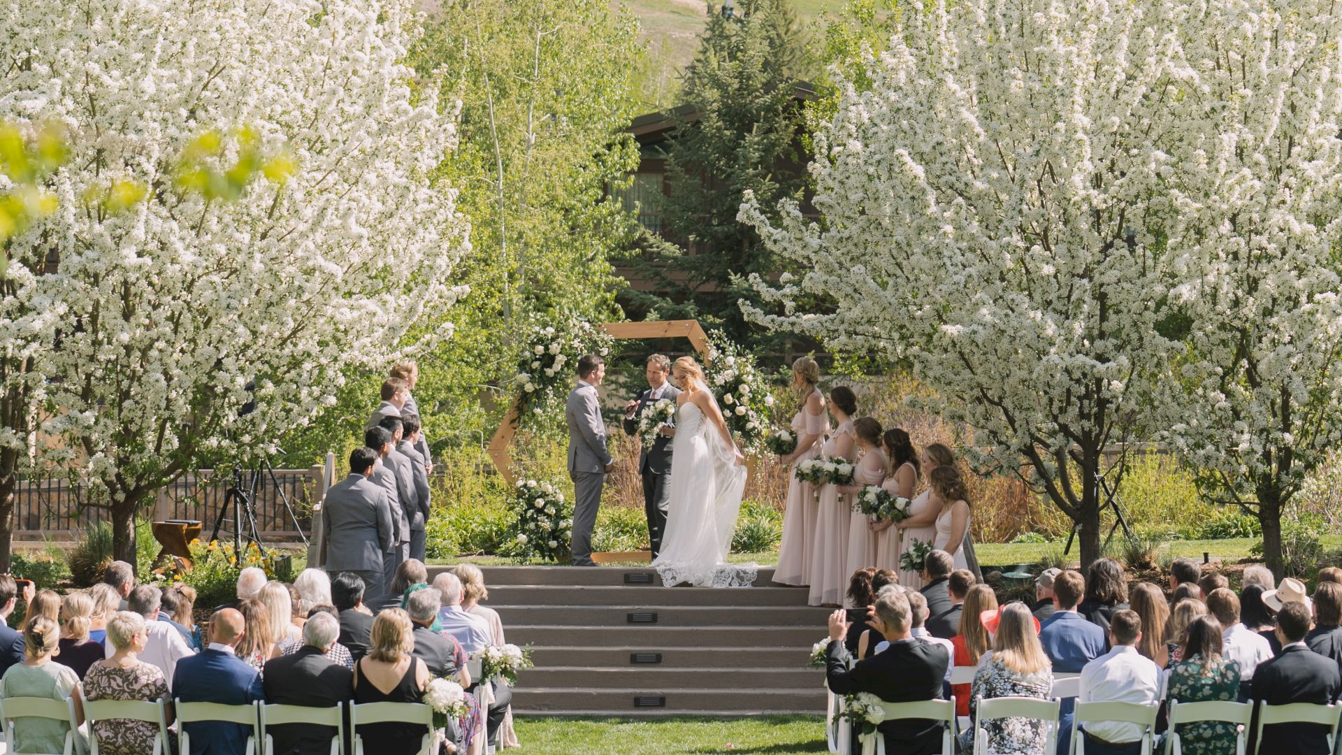 An outdoor wedding ceremony is taking place among blooming trees, with the bride and groom standing on a small stage with guests seated around them.