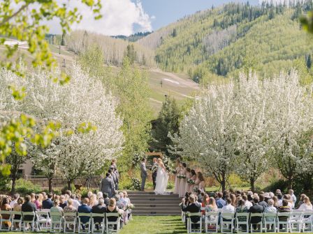 An outdoor wedding ceremony with guests seated on either side of an aisle, set against a backdrop of flowering trees and rolling hills.