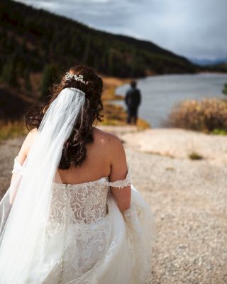 A bride in a wedding dress and veil is walking toward a figure standing by a lake, with mountains and cloudy skies in the background.