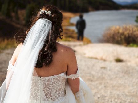 A bride in a wedding dress and veil is walking toward a figure standing by a lake, with mountains and cloudy skies in the background.
