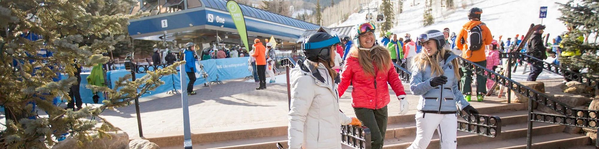 People in winter clothing at a ski resort, walking near a ski lift station with snow-covered mountains and trees in the background.