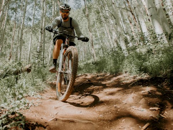 A person is mountain biking on a dirt trail surrounded by tall trees, wearing a helmet and sunglasses in a forest setting with dappled sunlight.
