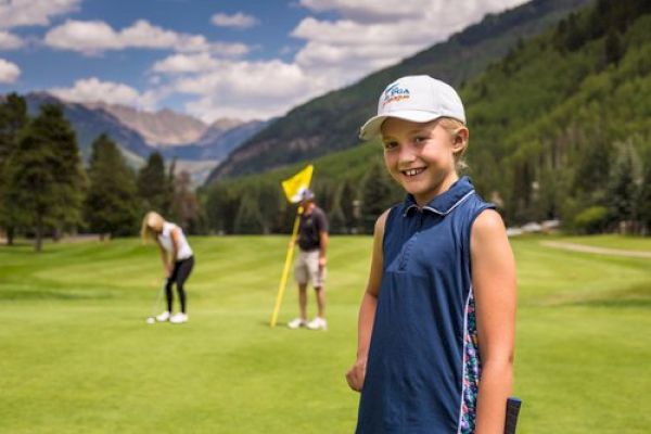 A young girl smiles in the foreground of a golf course with two people playing golf in the background, set against a mountainous landscape.