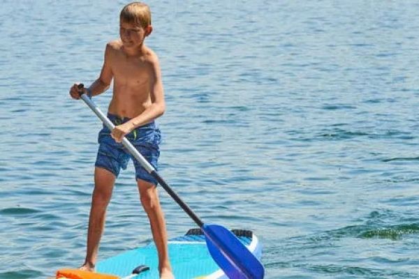 A shirtless boy is standing on a paddleboard in the water, using a paddle to navigate on a sunny day.