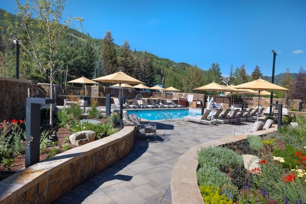 A serene outdoor pool area is surrounded by lounge chairs, umbrellas, and lush greenery against a backdrop of forested hills under a clear blue sky.