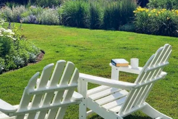 Two white Adirondack chairs sit on a green lawn, facing a garden. A small table between them holds a book and a white cup, hinting at relaxation.