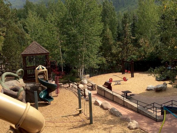 The image shows a playground with slides and climbing structures surrounded by trees and mountains in the background. The ground is covered with wood chips.