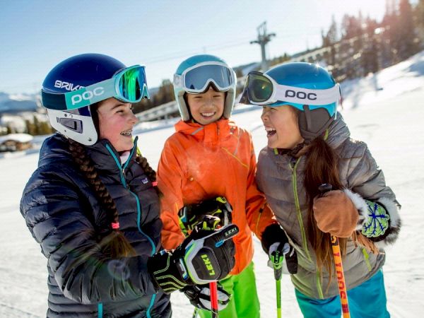 Three children dressed in ski gear and helmets are laughing and talking on a snowy slope on a sunny day.