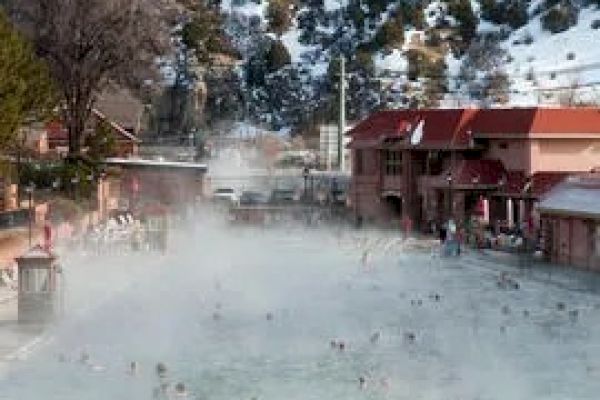 A steaming outdoor pool with people swimming, surrounded by snow-covered mountains and hills, alongside a building with red roofs and people walking nearby.