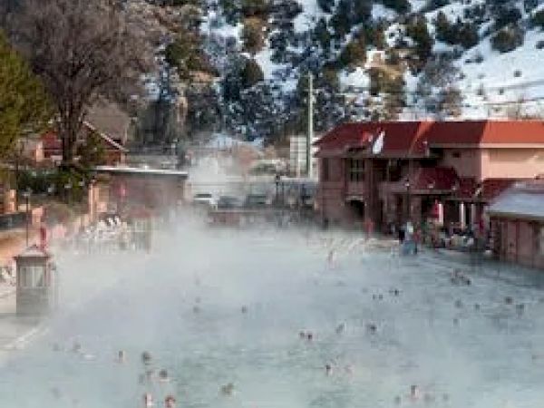 A steaming outdoor pool with people swimming, surrounded by snow-covered mountains and hills, alongside a building with red roofs and people walking nearby.