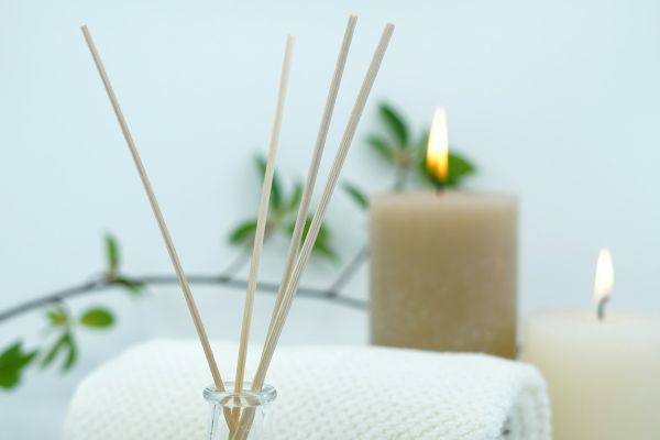 The image shows a spa setting with reed diffusers, a white rolled towel, two lit candles, and some greenery in the background.