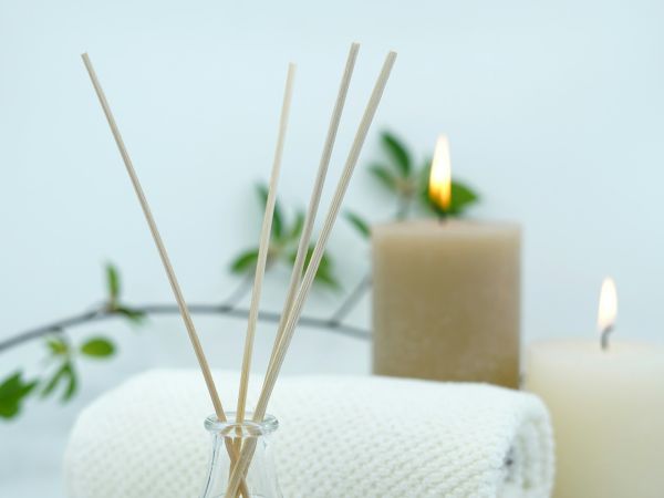 The image shows a spa setting with reed diffusers, a white rolled towel, two lit candles, and some greenery in the background.