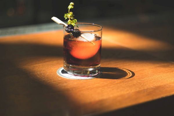 A cocktail is placed on a wooden table, garnished with a twist and berries, illuminated by a spotlight, creating a warm, inviting ambiance.