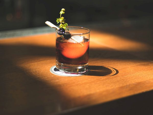 A cocktail is placed on a wooden table, garnished with a twist and berries, illuminated by a spotlight, creating a warm, inviting ambiance.