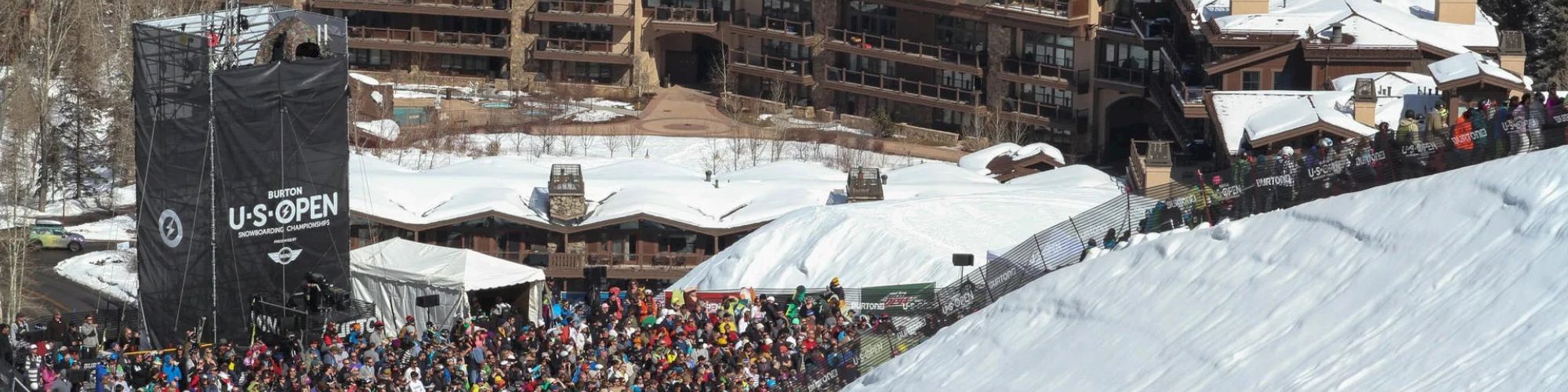 A large crowd gathers at a snowy resort with buildings in the background and a 