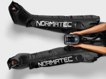 A person is using NormaTec compression boots, controlling the settings via a handheld device.