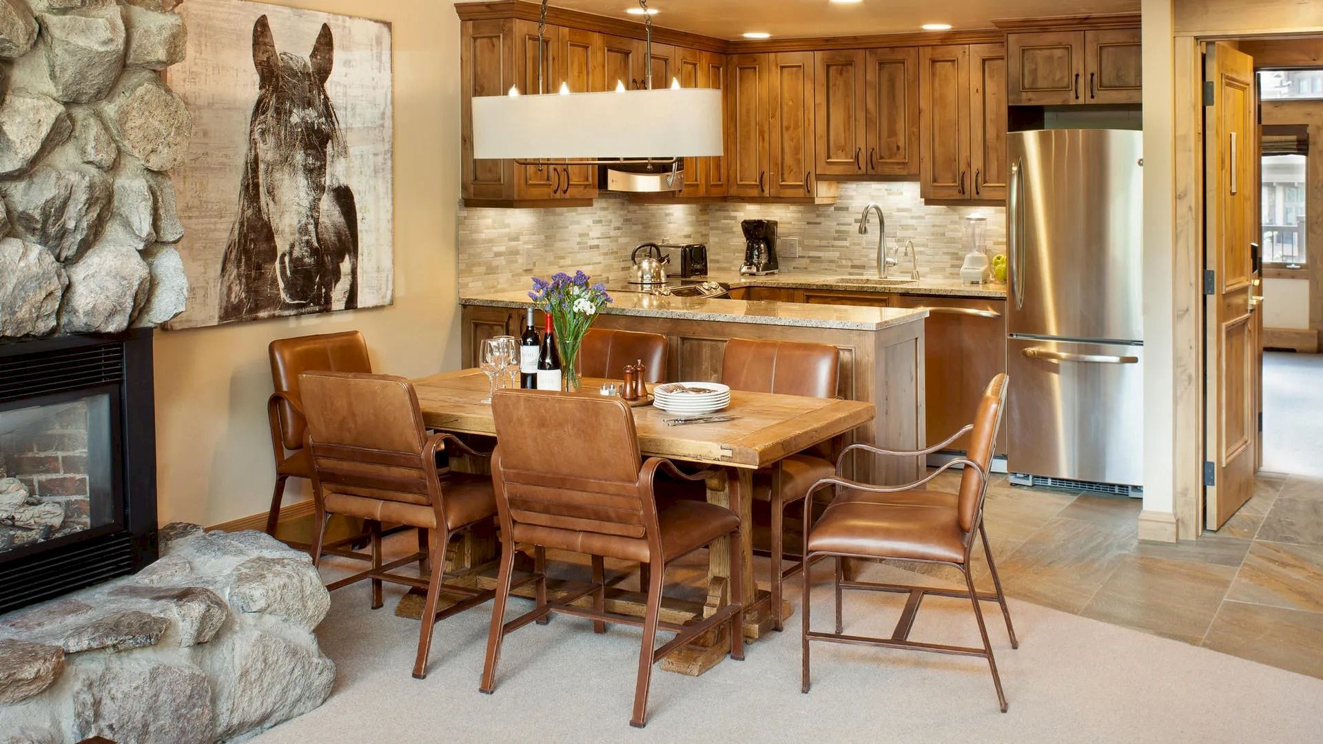 The image shows a rustic kitchen and dining area with wooden cabinets, a stone fireplace, leather chairs, and a horse painting on the wall.