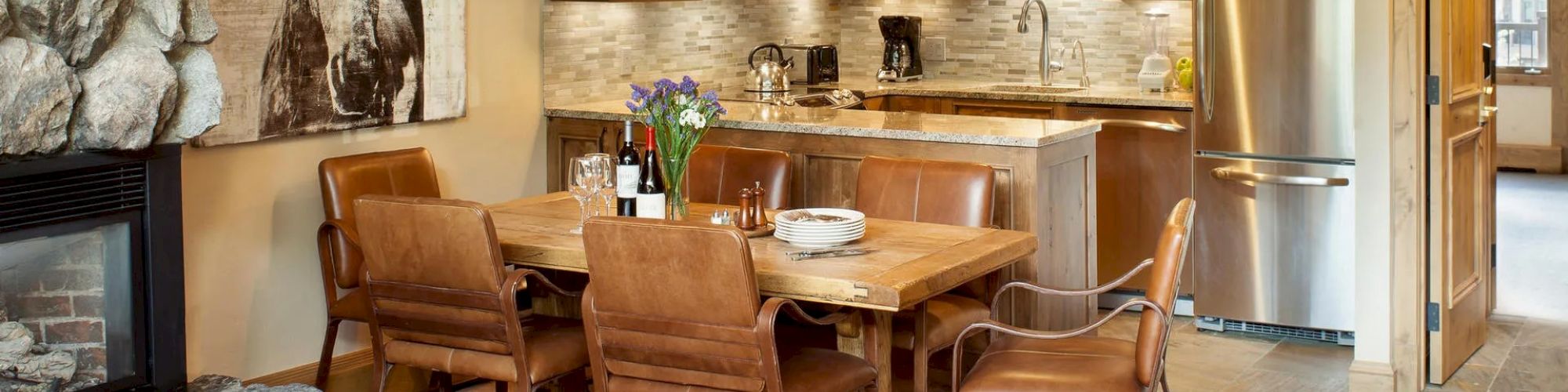 The image shows a rustic kitchen and dining area with wooden cabinets, a stone fireplace, leather chairs, and a horse painting on the wall.